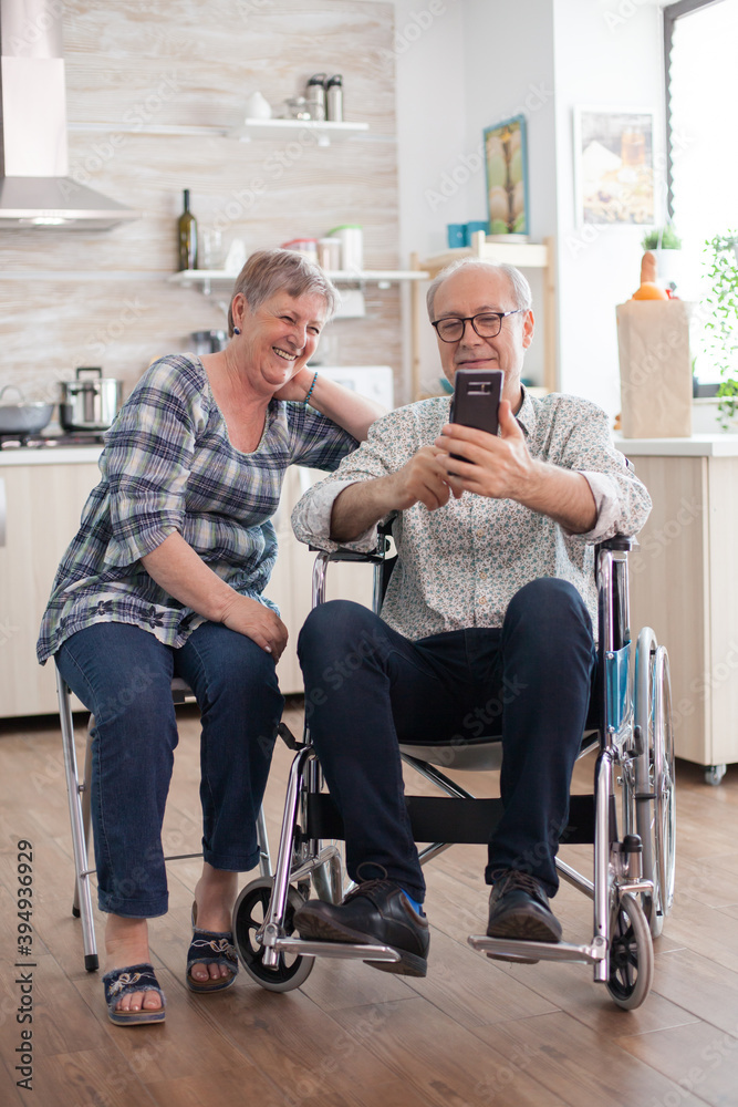 Disabled senior man in wheelchair and his wife Laughing and browsing on modern smartphone in kitchen. Paralyzed old man and his wife having a online conference.