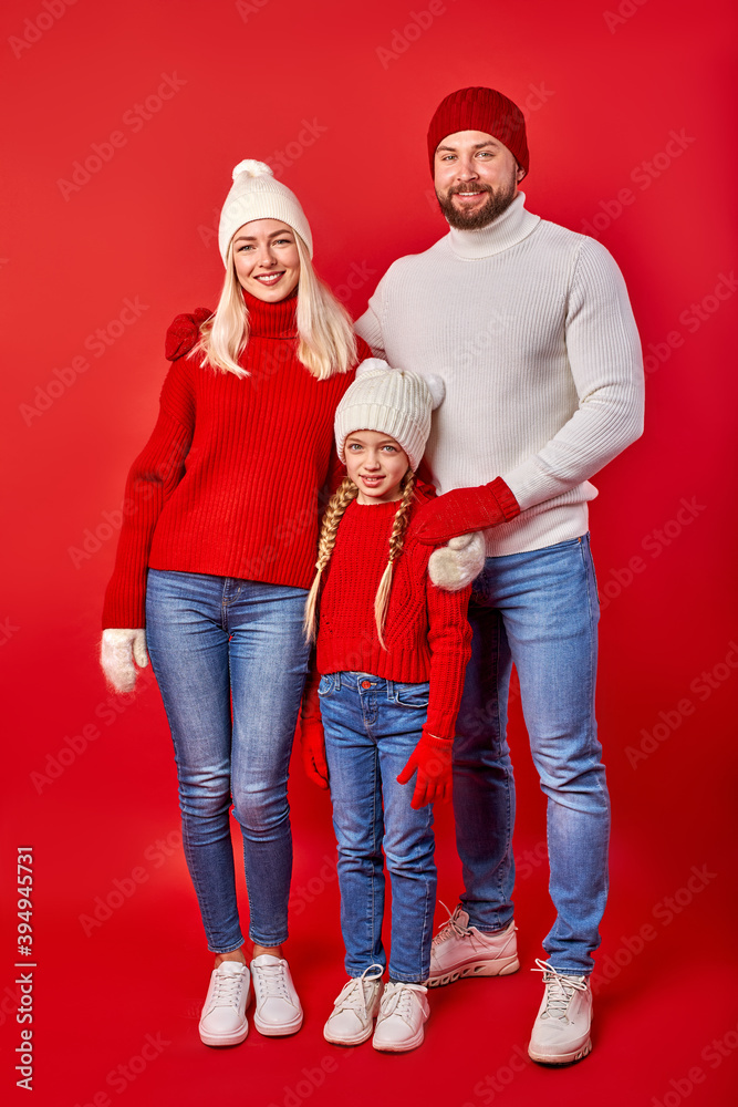 Fototapeta premium portrait of young parents with daughter in winter clothes, wearing sweaters, gloves and hats, looking at camera isolated over red background