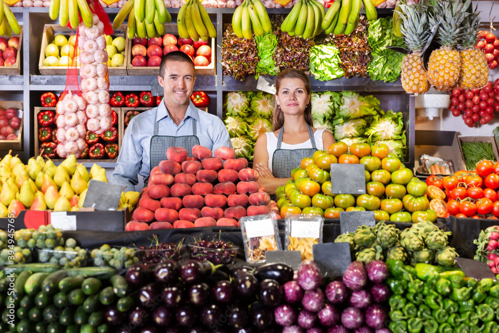 Young man and woman sellers in aprons standing near counter with a fresh vegetables and fruits at market