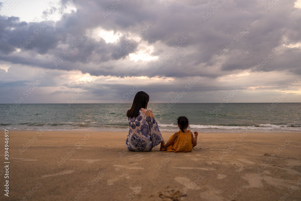 Young Mother and Daughter Sitting At Sea Shore Against Cloudy Sky at dusk.