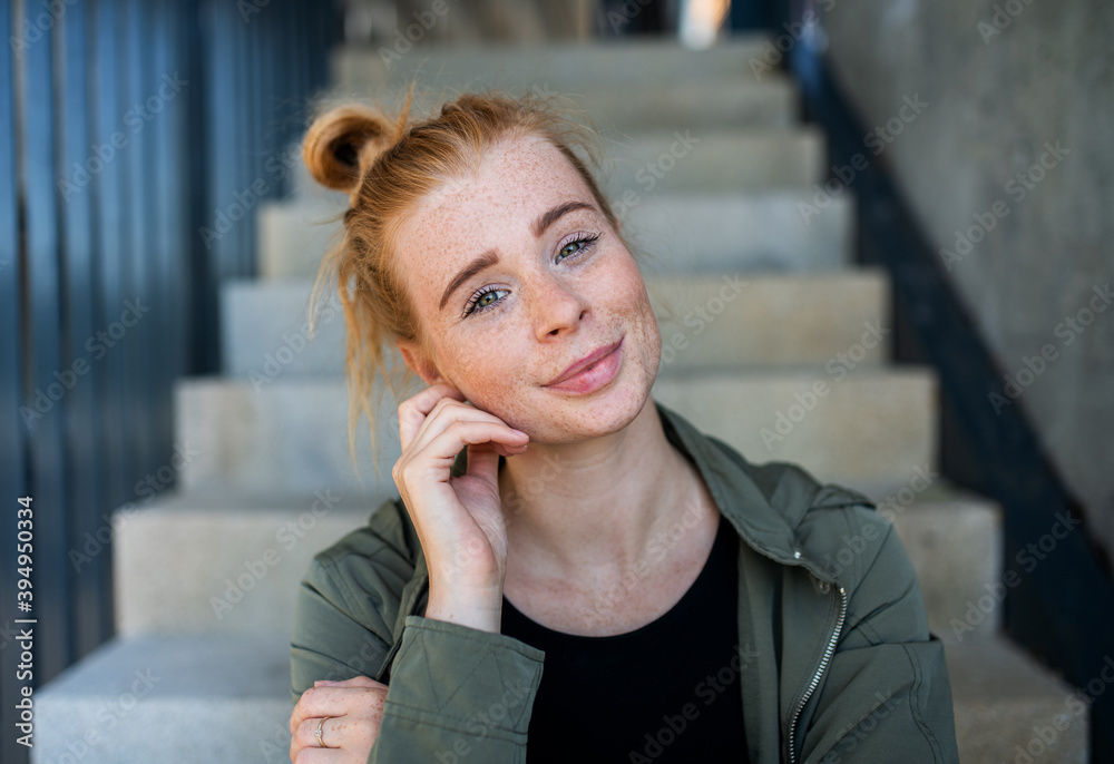Fototapeta premium Front view portrait of young woman with red hair outdoors in town, looking at camera.