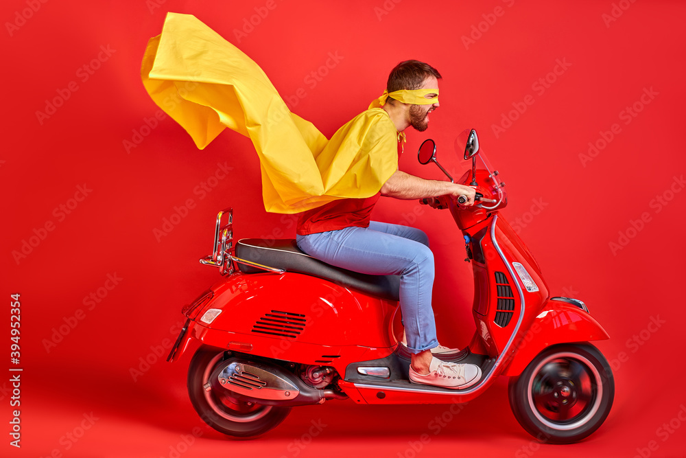 overjoyed mad crazy man driving moped fast speed wearing super hero costume isolated over red background, cloak sways in the wind overjoyed mad crazy man driving moped fast speed wearing super hero costume isolated over red background, cloak sways in the wind
