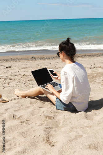 Young woman using laptop computer and holding a cell phone on a beach. Vacation lifestyle communication. Freelance work concept