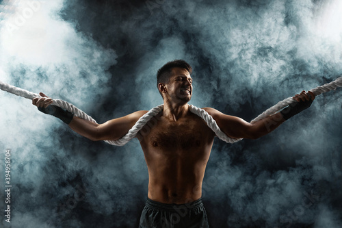 strong athletic muscular man with naked body holding rope on smoke background © producer