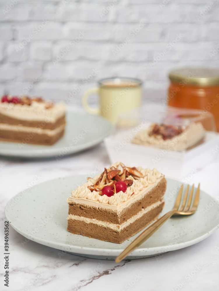 A piece of a light layered genoise sponge coffee cake topped with caramel almond in a plate with fork and coffee cup in background. It made with whole egg cake and butter A piece of a light layered genoise sponge coffee cake topped with caramel almond in a plate with fork and coffee cup in background. It made with whole egg cake and butter