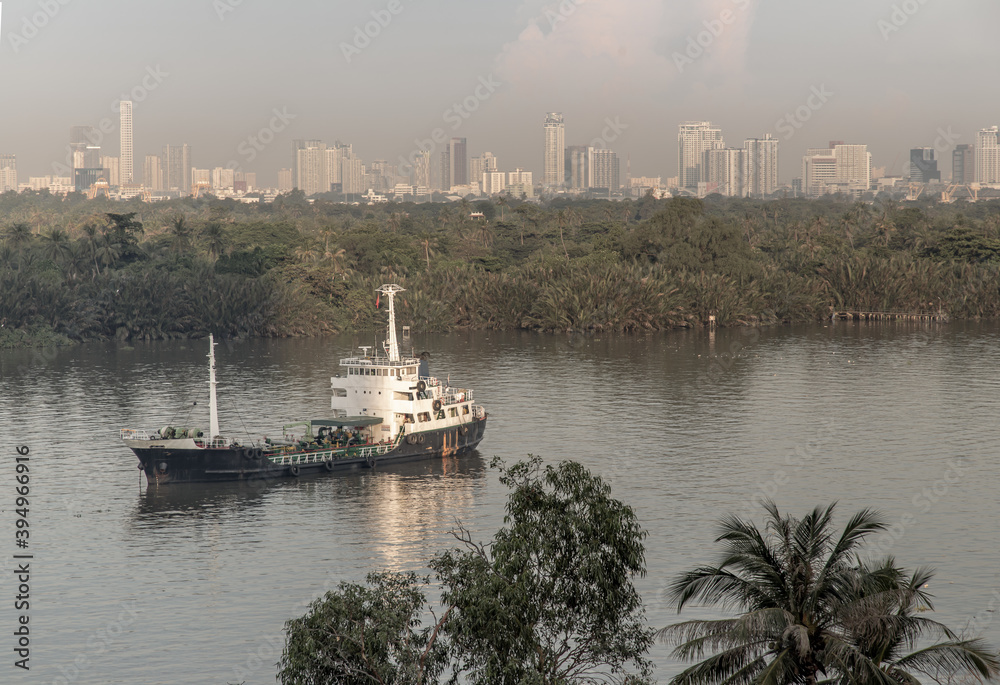 Cargo ship parked in the middle of the river and in front of a green forest with skyscrapers at evening by the Chao Phraya River.