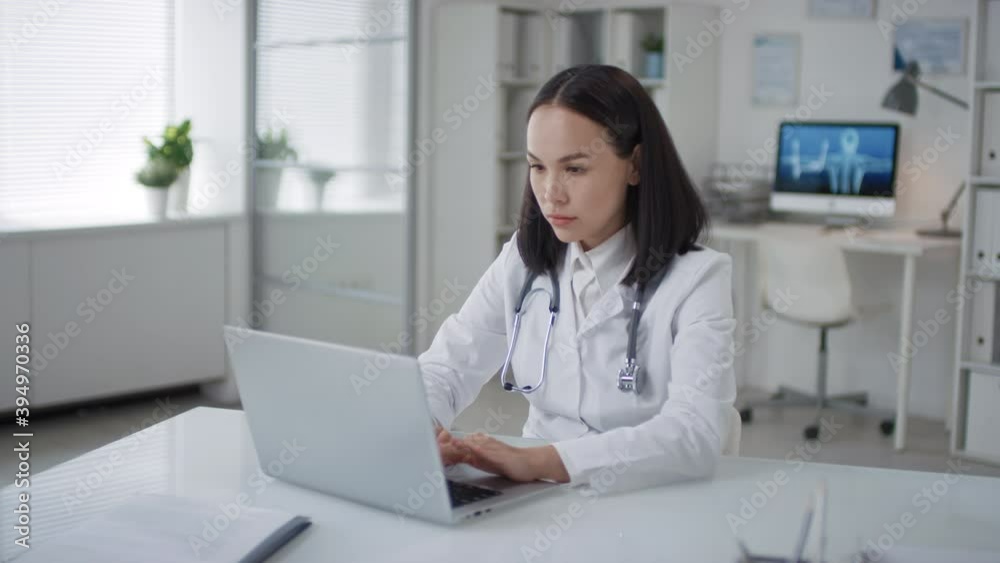 Medium shot portrait of Asian female doctor wearing white coat sitting at desk in her office room typing something on laptop Medium shot portrait of Asian female doctor wearing white coat sitting at desk in her office room typing something on laptop