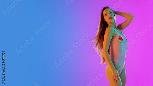 Tender. Beautiful seductive girl in fashionable pink swimsuit on gradient purple-blue background in neon light. Half-length portrait. Copyspace for ad. Summer, fashion, beauty, emotions concept.