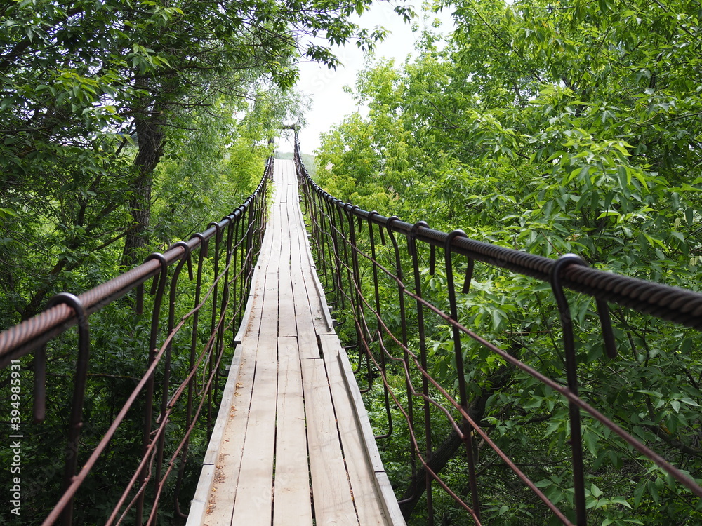 suspended wooden bridge over the river and meadow leading to the sky.