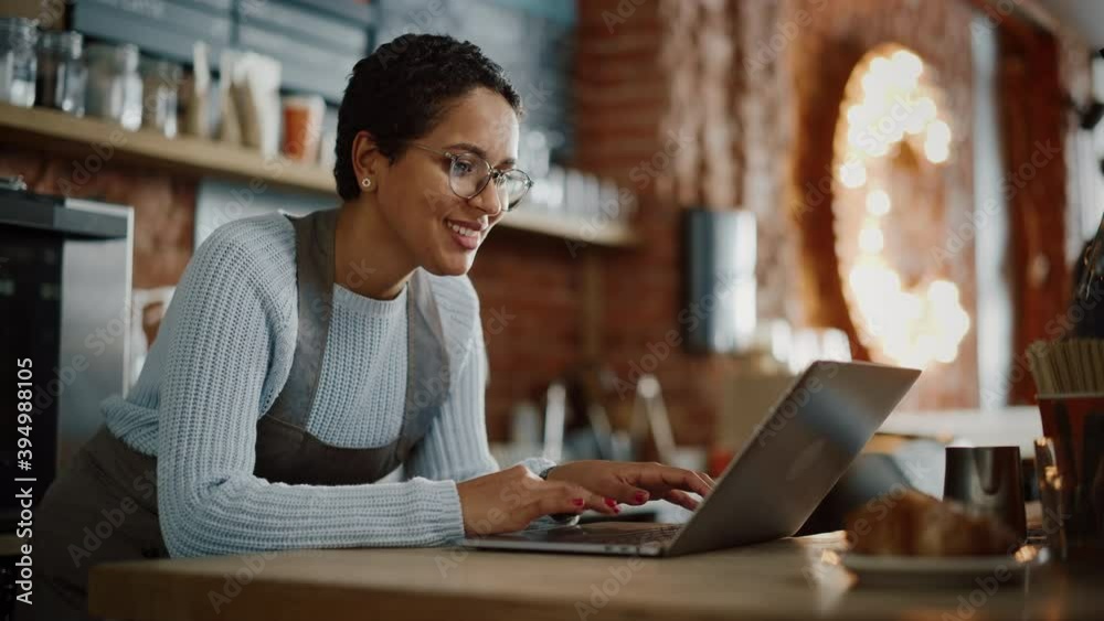 Young and Beautiful Latina Coffee Shop Owner is Working on Laptop Computer and Checking Inventory in a Cozy Cafe. Happy Restaurant Manager or Employee Browsing Internet and Chatting with Friends.