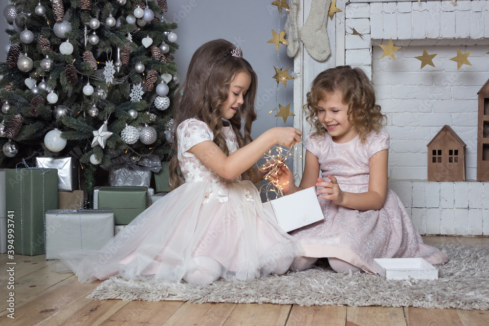 Two girls holding garland giftbox near fireplace. Christmas and new year decorated card. Sisters in beautiful gown sitting on the floor new year background