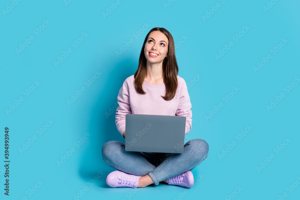 Full length photo of pretty lovely student girl sit floor crossed legs hold laptop look up empty space watch plane flying wear violet pullover sneakers denim isolated blue color background Full length photo of pretty lovely student girl sit floor crossed legs hold laptop look up empty space watch plane flying wear violet pullover sneakers denim isolated blue color background