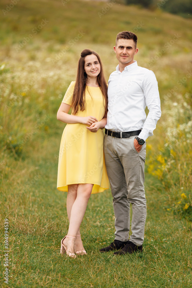 young happy couple in love in a grass field in sunny day. girl in a yellow dress and a guy with a stylish haircut. portrait of couple walking on countryside