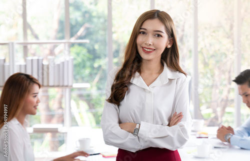 Young confident business woman standing in modern office. Portrait of young beautiful business woman in white in a busy modern workplace. Beautiful woman in office with business people.
