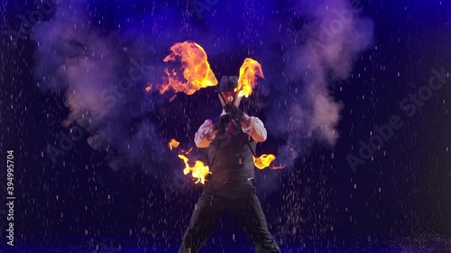 A professional circus performer rotates burning torches under streams of rain against the backdrop of a smoky studio with blue light. Close up in slow motion.
