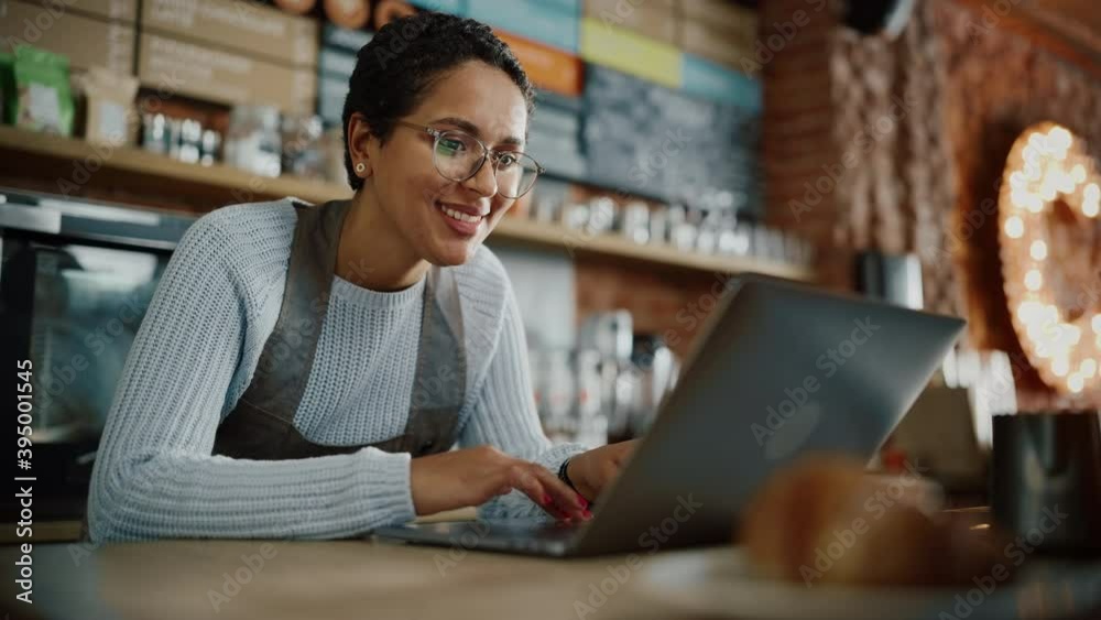 Young and Beautiful Latina Coffee Shop Owner is Working on Laptop Computer and Checking Inventory in a Cozy Cafe. Happy Restaurant Manager or Employee Browsing Internet and Chatting with Friends.