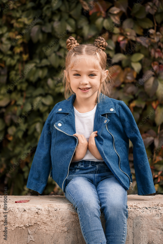 A beautiful stylish blonde girl in a blue jacket sits on a concrete block. Girl 7 years old with a smile on a background of leaves A beautiful stylish blonde girl in a blue jacket sits on a concrete block. Girl 7 years old with a smile on a background of leaves
