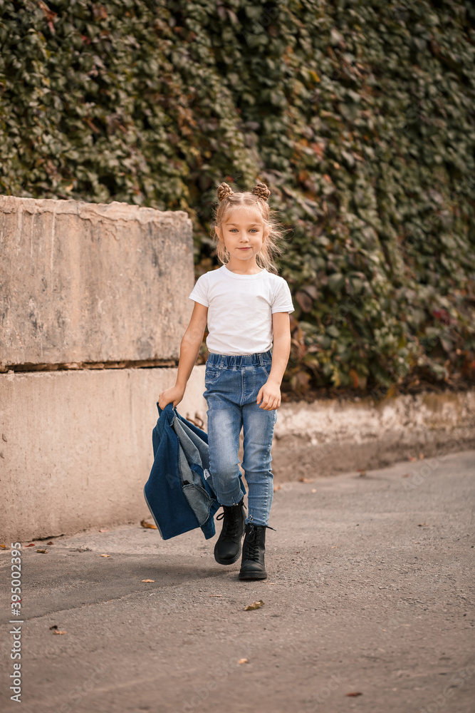 Stylish blonde little girl in jeans and a white T-shirt walks down the street. Girl 7 years old small model, beautiful child Stylish blonde little girl in jeans and a white T-shirt walks down the street. Girl 7 years old small model, beautiful child