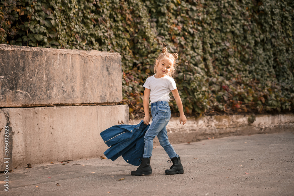 Stylish blonde little girl in jeans and a white T-shirt walks down the street. Girl 7 years old small model, beautiful child Stylish blonde little girl in jeans and a white T-shirt walks down the street. Girl 7 years old small model, beautiful child