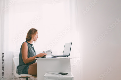 A young woman sitting at a table and leafing through a book. Nearby are a laptop and office supplies. Interior is white color. Copy space