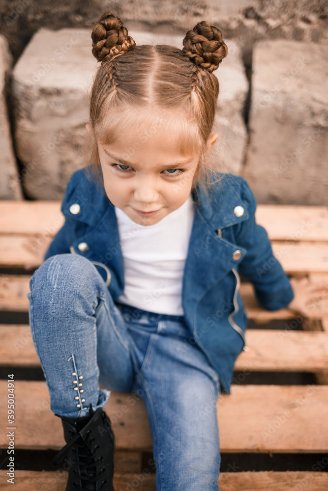 A beautiful little girl is sitting in stylish clothes on wooden pallets. Stylish child in a blue jacket. Casual style, fashion for children, fashionable suit, happy childhood A beautiful little girl is sitting in stylish clothes on wooden pallets. Stylish child in a blue jacket. Casual style, fashion for children, fashionable suit, happy childhood