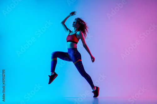 Run and jump. Young sportive woman training isolated on gradient blue-pink background in neon light. Athletic and graceful. Modern sport, action, motion, youth concept. Beautiful female practicing.