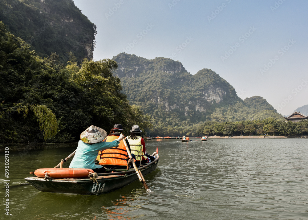 Tourists on a boat tour in Trang An Landscape Complex. There is a lady wearing a vietnamese traditional hat rowing. Ninh Binh, Vietnam.