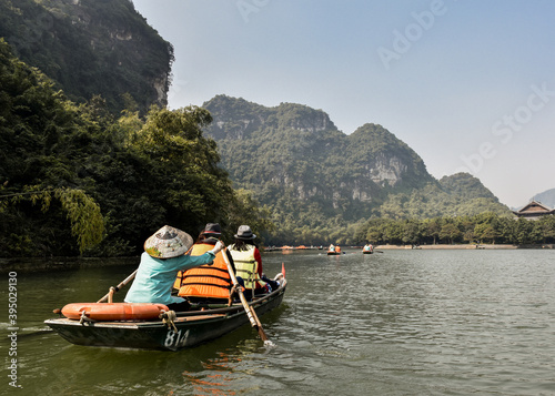 Tourists on a boat tour in Trang An Landscape Complex. There is a lady wearing a vietnamese traditional hat rowing. Ninh Binh, Vietnam.