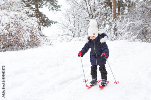 Children's feet in red plastic skis with sticks go through the snow from a slide-a winter sport, family entertainment in the open air. A little girl glides down the slope from an early age. Copy space