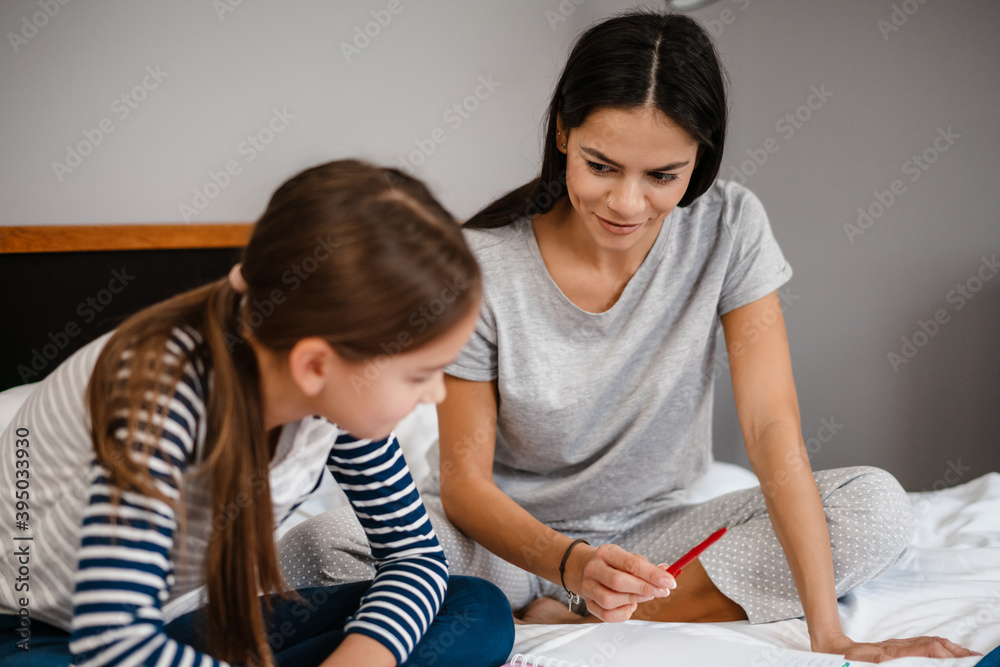 Nice beautiful mother and daughter doing homework while sitting on bed Nice beautiful mother and daughter doing homework while sitting on bed