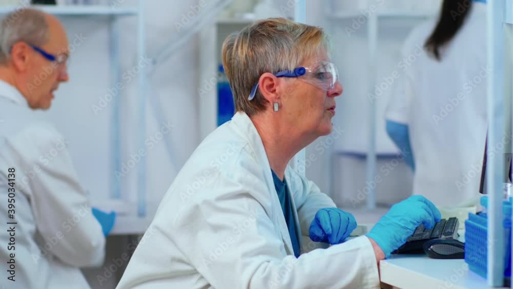 Close up of elderly chemist woman typing on computer in modern equipped laboratory. Multiethnic stuff analysing vaccine evolution using high tech for researching treatment against covid19 virus Close up of elderly chemist woman typing on computer in modern equipped laboratory. Multiethnic stuff analysing vaccine evolution using high tech for researching treatment against covid19 virus
