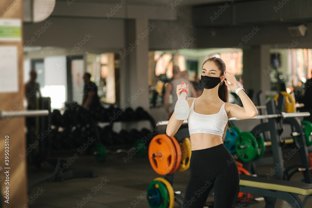 Brunettr woman with face mask cleaning exercise machine with disinfectant. Woman exercising in a gym during coronavirus epidemic
