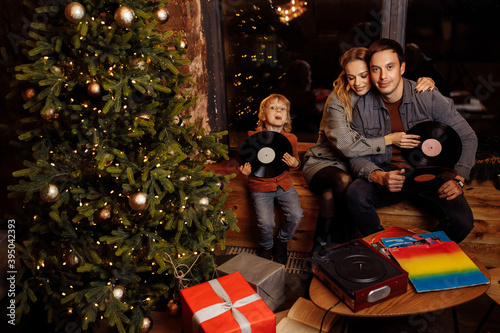 Happy parents with little son sit near Christmas tree, happy family listening music plates, smiling, enjoy festive mood, vinyl music, winter holidays at home, xmas concept