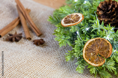 close-up of scattered slices of dry orange on green wreath with lights and cone, cinnamon sticks with anise on jute background, christmas rustic vintage decoration, exotic raw fruit, front view