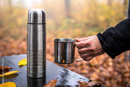 Woman holding cup, thermos with hot drink on table. Refreshment during hike in autumn woodland