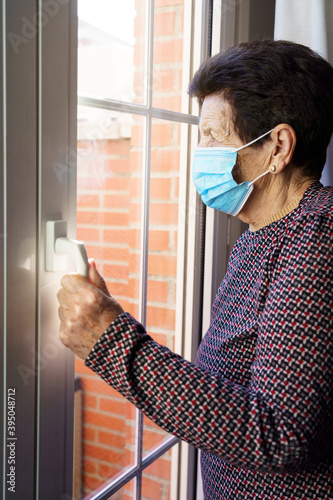 Mujer blanca de noventa años con mascarilla en confinamiento por coronavirus o covid-19, en el interior de su casa, mirando por la ventana. 