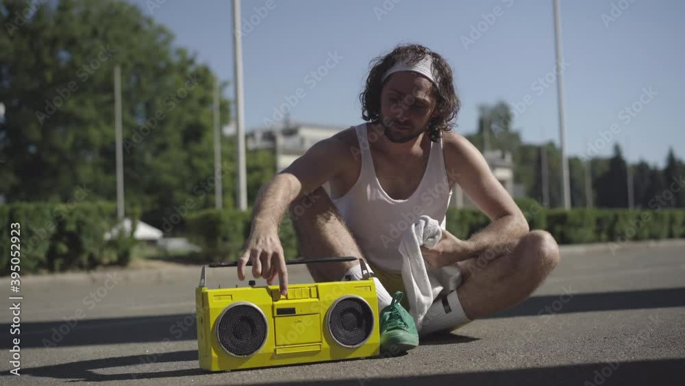Exhausted young sportsman wiping face with white towel, turning off retro tape recorder, and smiling at camera. Portrait of handsome satisfied 1980s Caucasian man after training outdoors.
