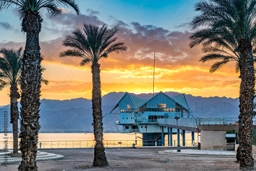 Colorful sunrise above mountains and public beach with promenade and decorative palms as foreground, Red Sea, Middle East