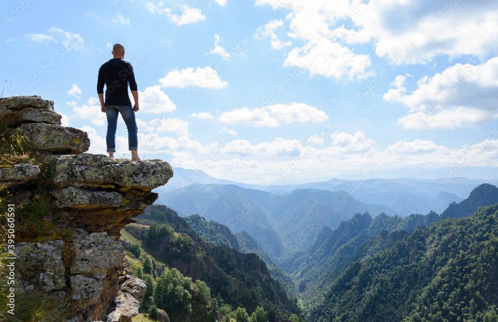 A young brutal man stands on the edge of a mountain cliff and meditates, enjoying the view of mountain peaks.