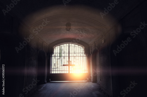 Vaulted arch in an old house with closed iron gates. On the grate of the crossbar in the form of a cross, the rays of the setting sun break through the fence