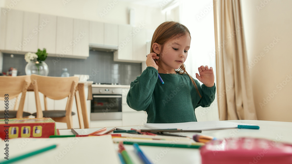 A small cute girl thinking about writing sitting at a table in a bright spacy kitchen A small cute girl thinking about writing sitting at a table in a bright spacy kitchen