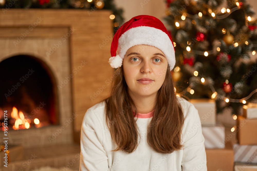 Christmas woman in santa hat sitting in decorating living room near fireplace and fir tree, looks at camera, wearing casual shirt, has calm facial expression. Christmas woman in santa hat sitting in decorating living room near fireplace and fir tree, looks at camera, wearing casual shirt, has calm facial expression.