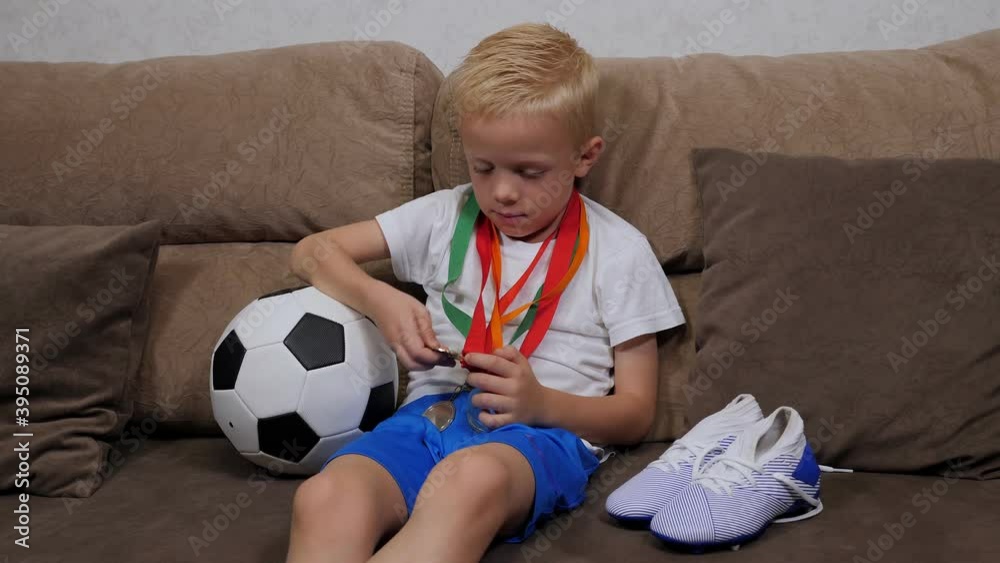 A little boy soccer player is sitting on the couch at home with a soccer ball, soccer cleats and lots of medals. Football champion. Children's achievements. A little boy soccer player is sitting on the couch at home with a soccer ball, soccer cleats and lots of medals. Football champion. Children's achievements.