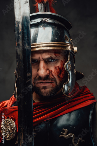 Serious and angry roman soldier dressed in dark armour with helmet and red mantle posing looking at camera and holding a sword.