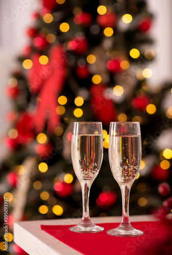 Two flute glasses with sparkling drink (champagne) standing on table covered with red tablecloth. Blurred decorated Christmas tree and lights in background. Winter advent holidays concept