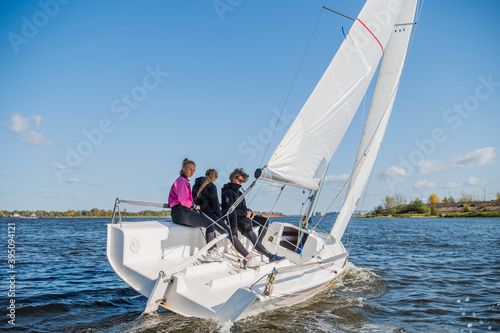 A beautiful white racing single-masted yacht is sailing against a beautiful river landscape with a blue sky. A man and two girls are on board