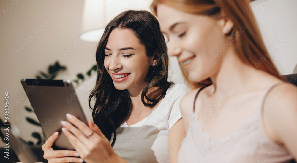 Charming caucasian lady with red hair and freckles near her brunette friend are using a tablet and smile Charming caucasian lady with red hair and freckles near her brunette friend are using a tablet and smile