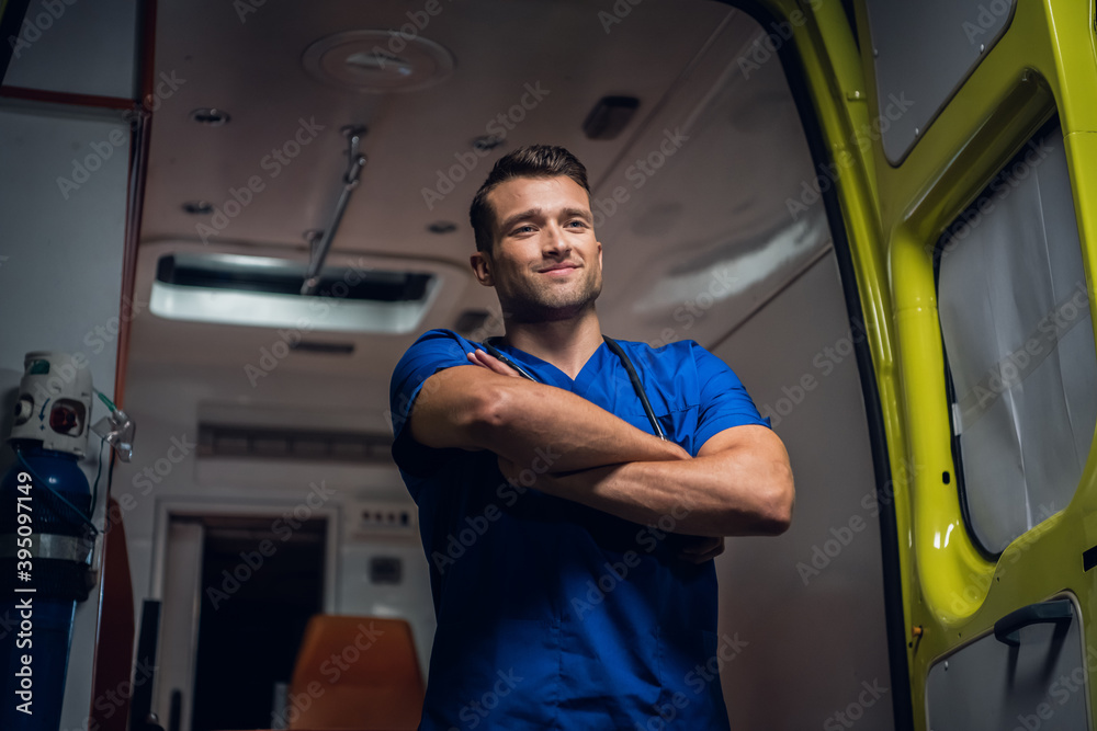 A portrait of a young intern standing in front of an ambulance car with his hands folded and smiling. A portrait of a young intern standing in front of an ambulance car with his hands folded and smiling.
