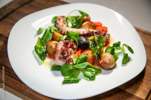 A plate of a healthy and delicious grilled ostopus, potatoes, black olives, spinach leaves and tomatoe sauce (on a wooden board)