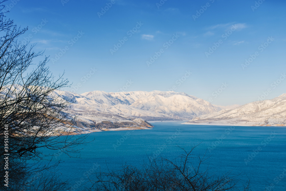 Charvak reservoir with blue water on a clear winter day in Uzbekistan, surrounded by snow-capped mountain peaks of Tien Shan